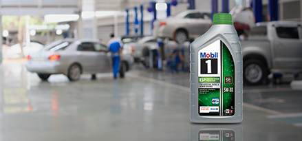 A one litre bottle of Mobil 1 5W-30 motor oil displayed in a car repair shop with vehicles and a mechanic in the blurred background.